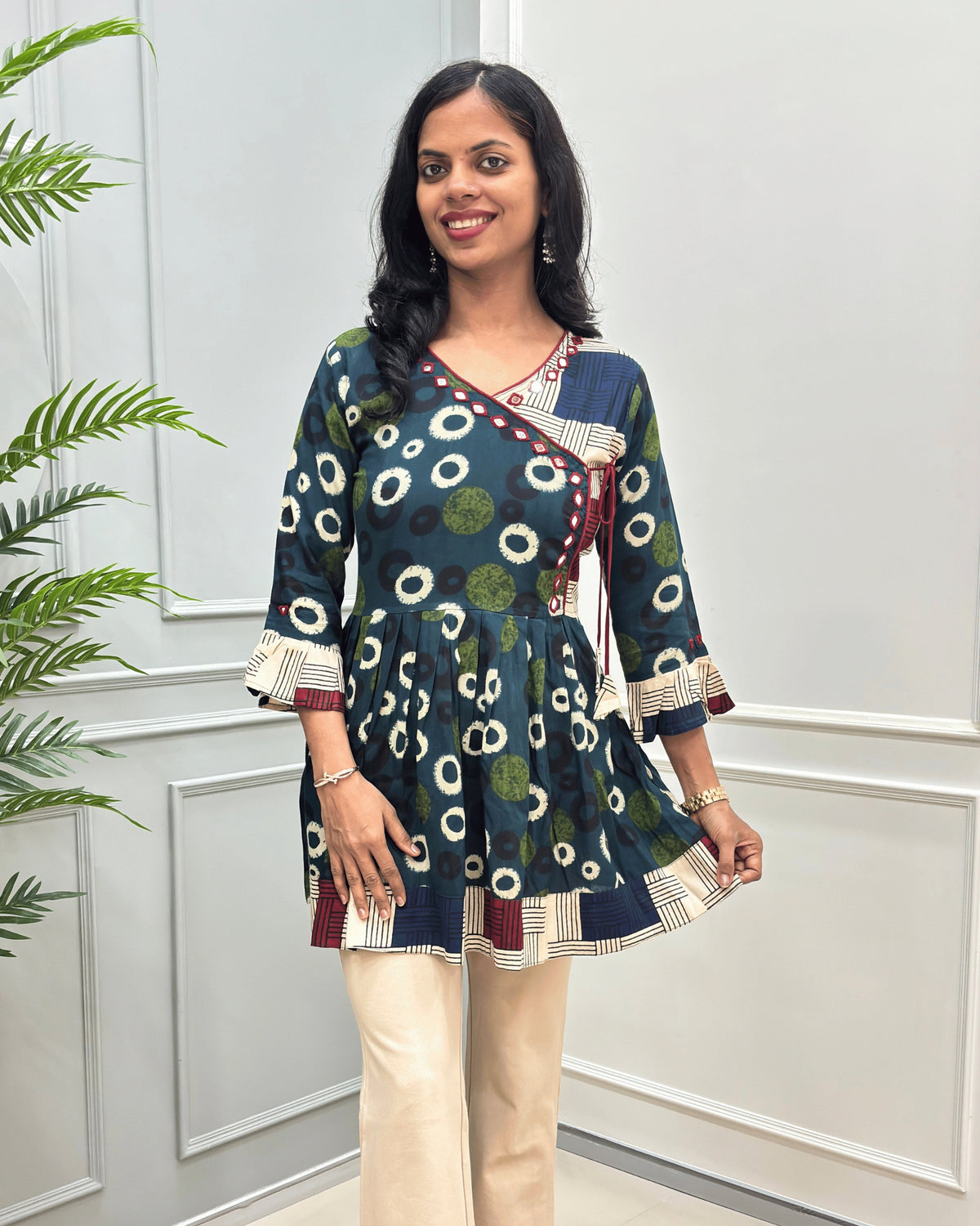 Front view of Green women's Ajrakh Plant based cotton Short-Kurti with a multicolor abstract print of circles and geometric patterns, Beige Strip print on top bottom & sleeves cuffs in paired with beige pants, standing in front of a white paneled wall.
