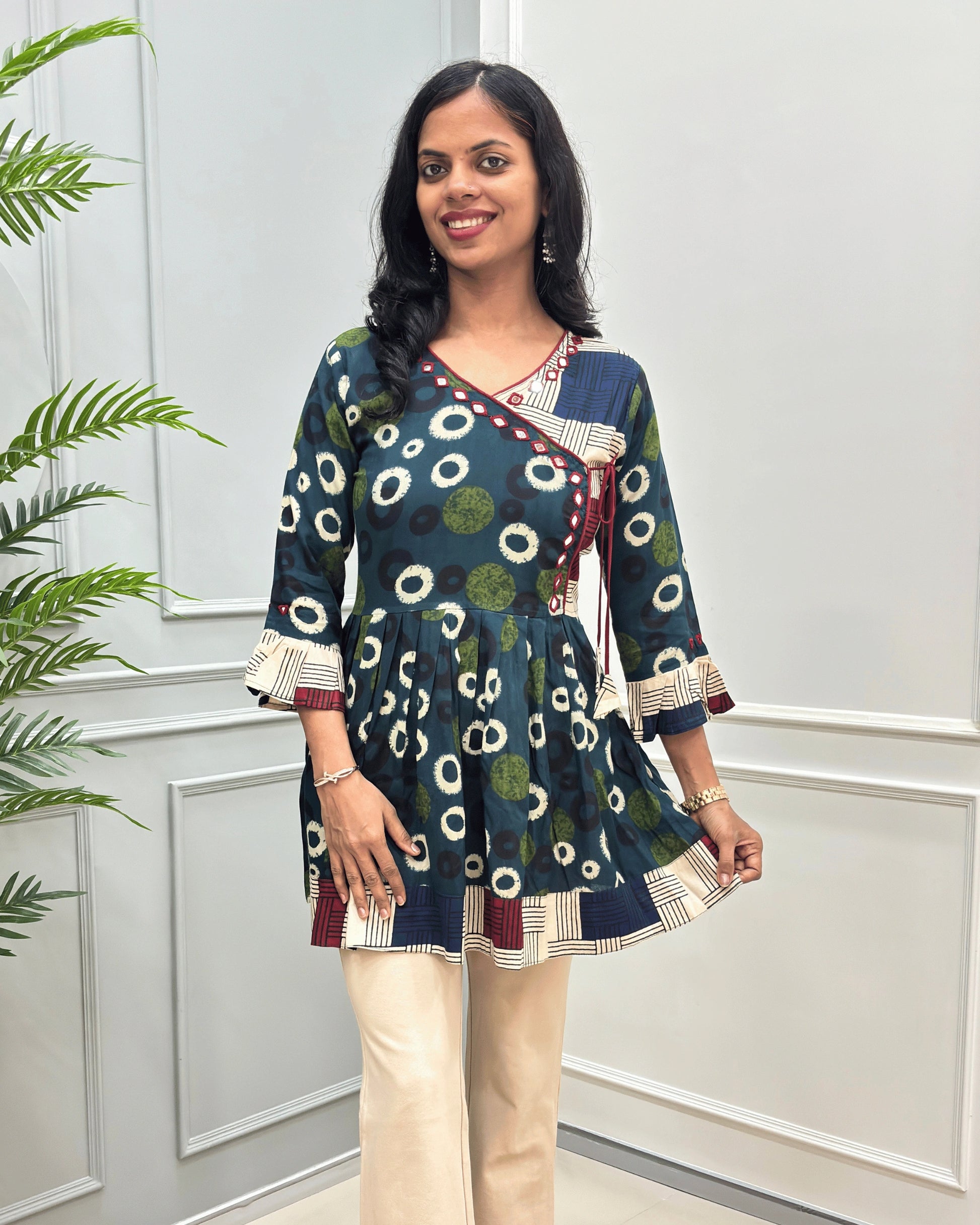 Front view of Green women's Ajrakh Plant based cotton Short-Kurti with a multicolor abstract print of circles and geometric patterns, Beige Strip print on top bottom & sleeves cuffs in paired with beige pants, standing in front of a white paneled wall.