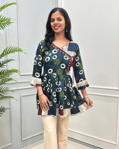 Front view of Green women's Ajrakh Plant based cotton Short-Kurti with a multicolor abstract print of circles and geometric patterns, Beige Strip print on top bottom & sleeves cuffs in paired with beige pants, standing in front of a white paneled wall.