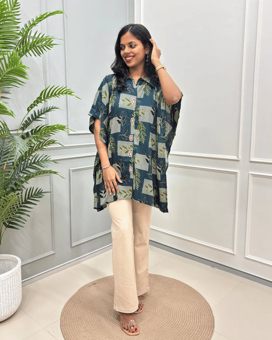 Green Women's Ajrakh Plant Based Rayon Cotton Short Kaftan with a Green and Deep Green Leaf print along with beige botanical block print, paired with beige wide-flared pant, standing on a round woven rug in front of a white paneled wall.