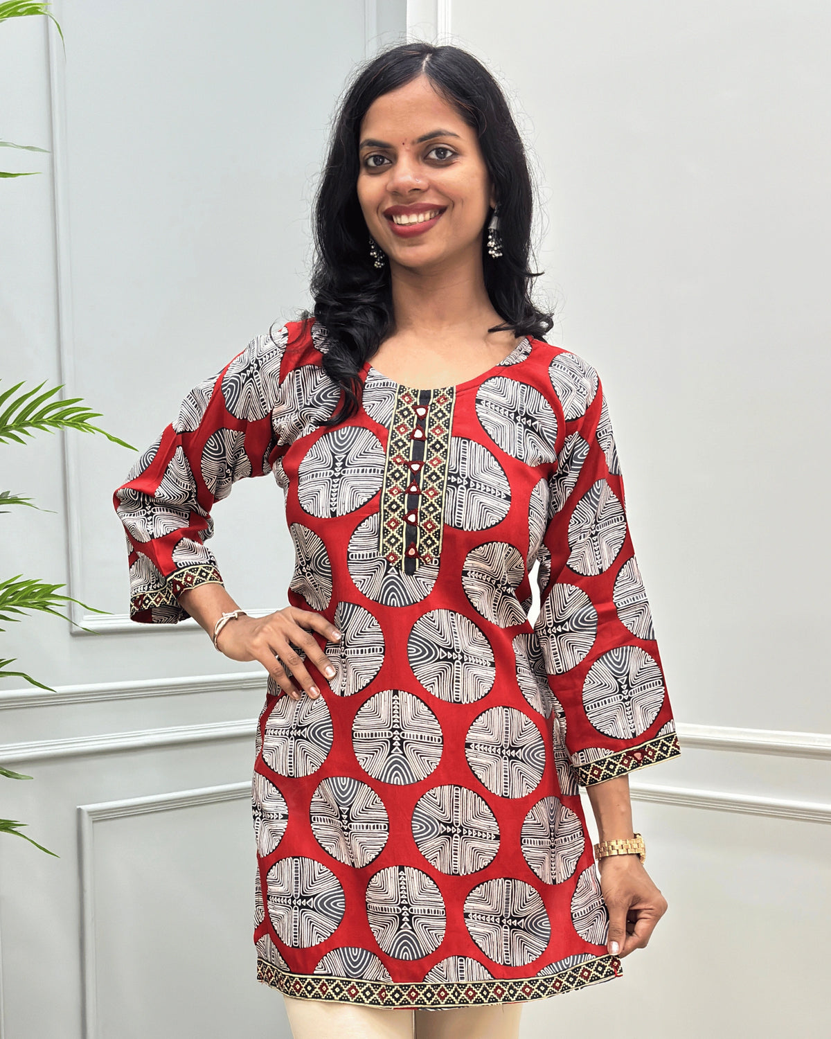 Close-up of Red women's plant based cotton short kurti with a Black , and white circular geometric pattern, Ajrakh print & Hand-mirror work on front and sleeves. standing against a light gray paneled wall. 