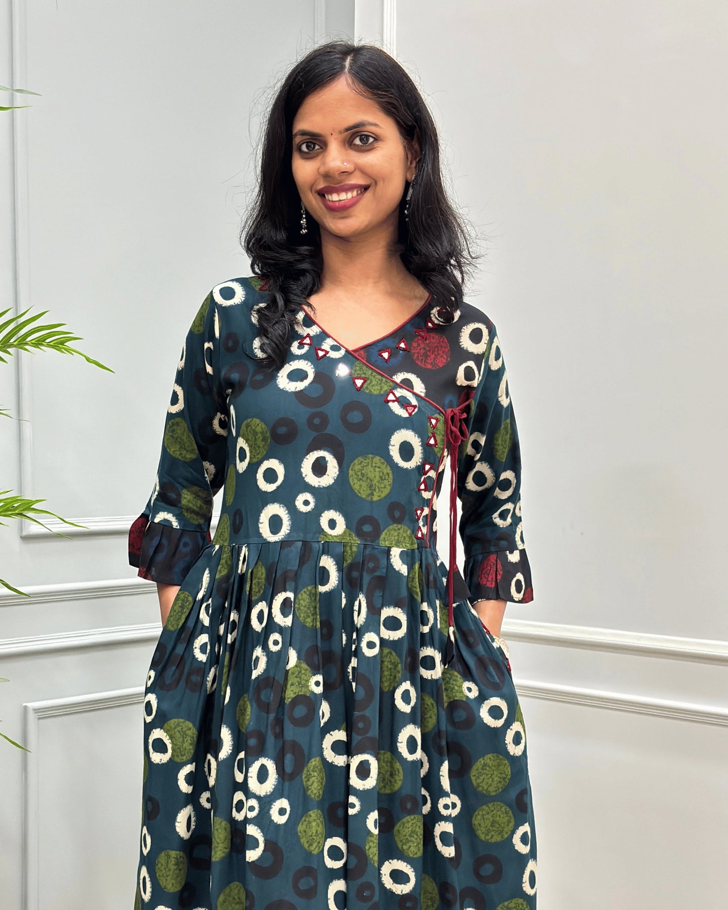 Close-up of Green women's plant based cotton A-line Long-kurti with a green, white, and black abstract circle print, three quarter sleeves with frill and red &white circle on black base. featured with Angrakha pattern along with pockets and hand-mirror work on V-neck , standing against a white paneled 
