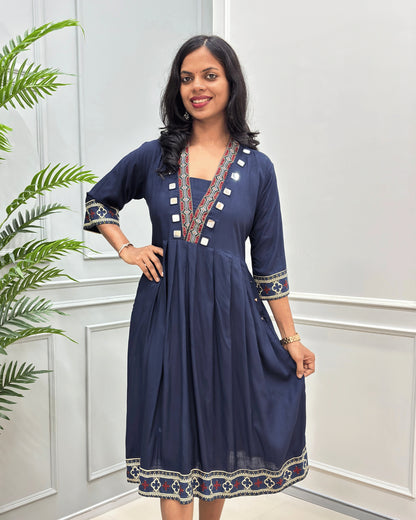  Navy-Blue women's plant based  cotton A-line long kurti dress with colorful geometric embroidery on the neckline, cuffs, and hem, standing in front of a white paneled wall with a green plant on the left.