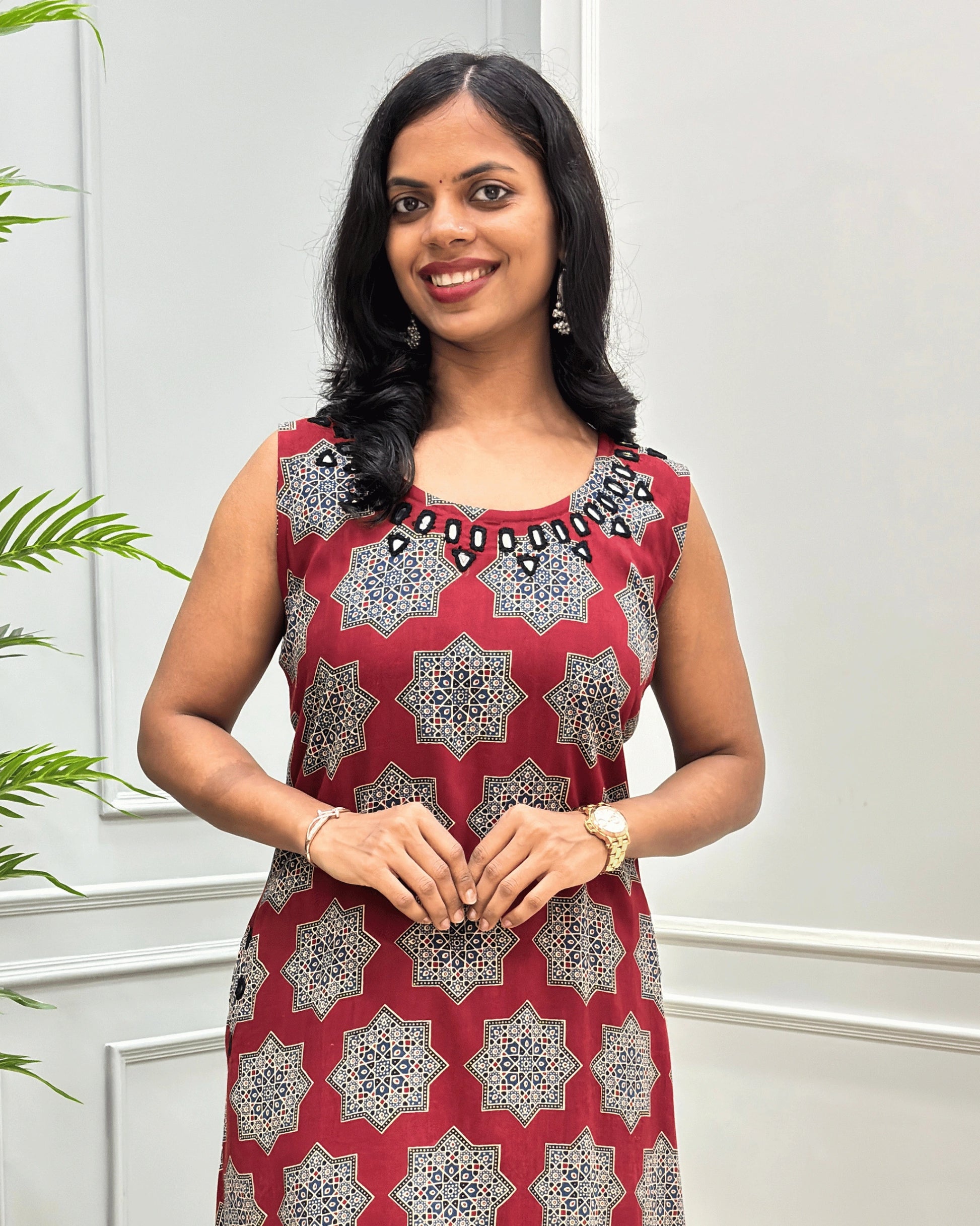 Close-up of Red cotton Long sleeveless Kurti with a geometric Black, Beige, and white starburst pattern, hand mirror work on neck border, standing against a light gray paneled wall.