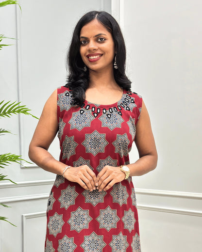 Close-up of Red cotton Long sleeveless Kurti with a geometric Black, Beige, and white starburst pattern, hand mirror work on neck border, standing against a light gray paneled wall.