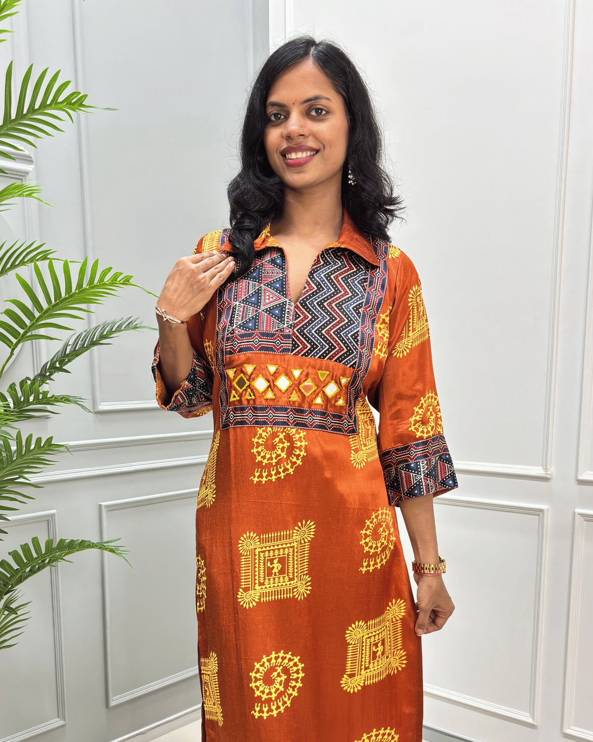 Close-up of Orange Ajrakh Mashru silk Long kurti with multicolor geometric , warli print on front & sleeves along eith hand -mirror work on front , Collar V shaped neck patterns, standing in front of a white paneled wall with a green plant on the left.