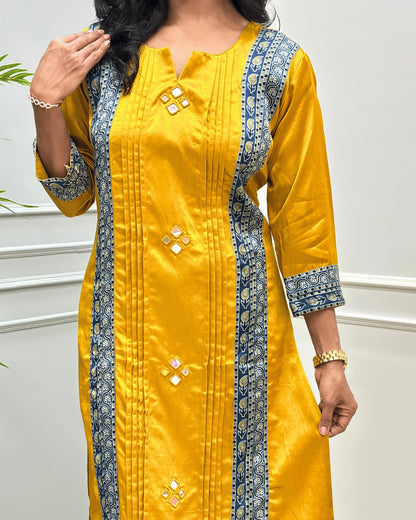 Close up of Yellow women's Ajrakh Mashru Silk Long-kurti with blue patterned borders and small mirror embellishments, shown from the shoulders to below the knees against a light indoor wall.