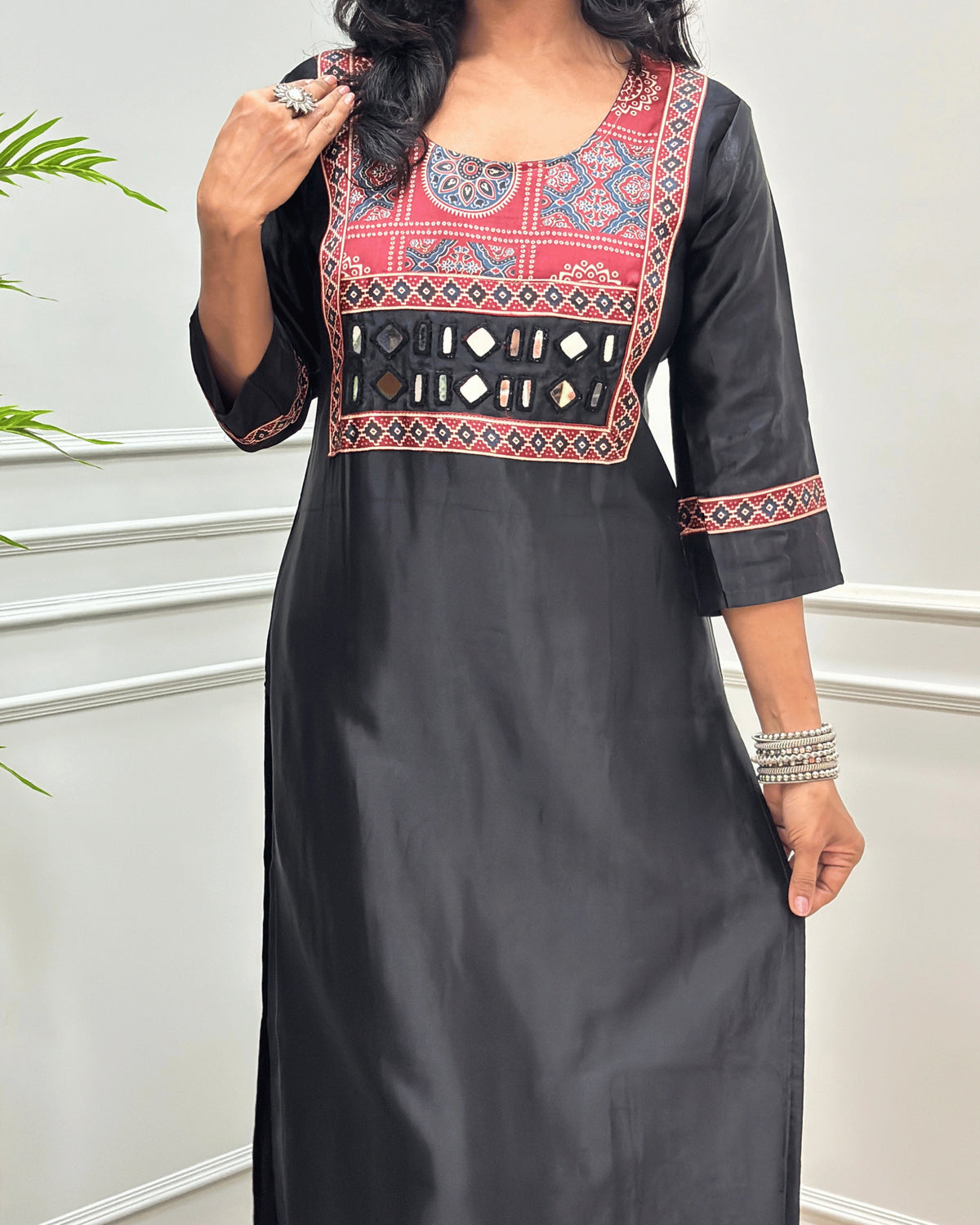 Close-up of Black Ajrakh Mashru Silk Long-kurti with a highly detailed Ajrakh geometric embroidered yoke in blue, red, and white, along with ajrakh print border on front & sleeves, posed against a light gray paneled wall.