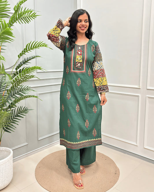 Green Women's Ajrakh Plant Based Rayon Cotton Co-Ord Set with colorful printed sleeves and embroidered details, standing on a round woven mat in front of a white paneled wall with a potted plant.