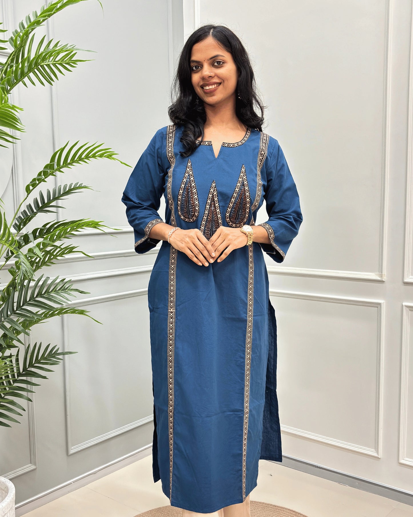 Front-view of Blue Women's Hand-block Ajrakh Pure Cotton Long Kurti with Beige and Black embroidered details on the V-neckline and borders on front & Sleeves , standing in a decorated room with a potted plant.