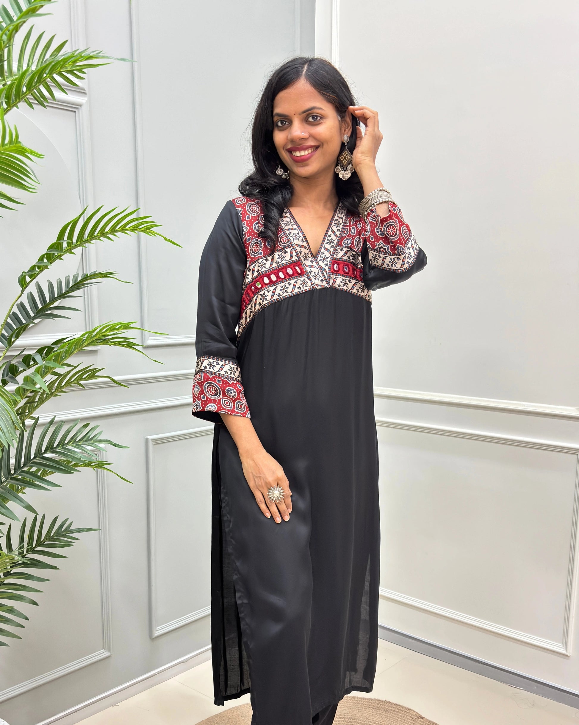 Front view of Black women's Ajrakh Modal Silk Long kurti featured with Black base & ajrakh print on red & white neck & cuffs , hand-mirror work on chest standing in a white paneled room.