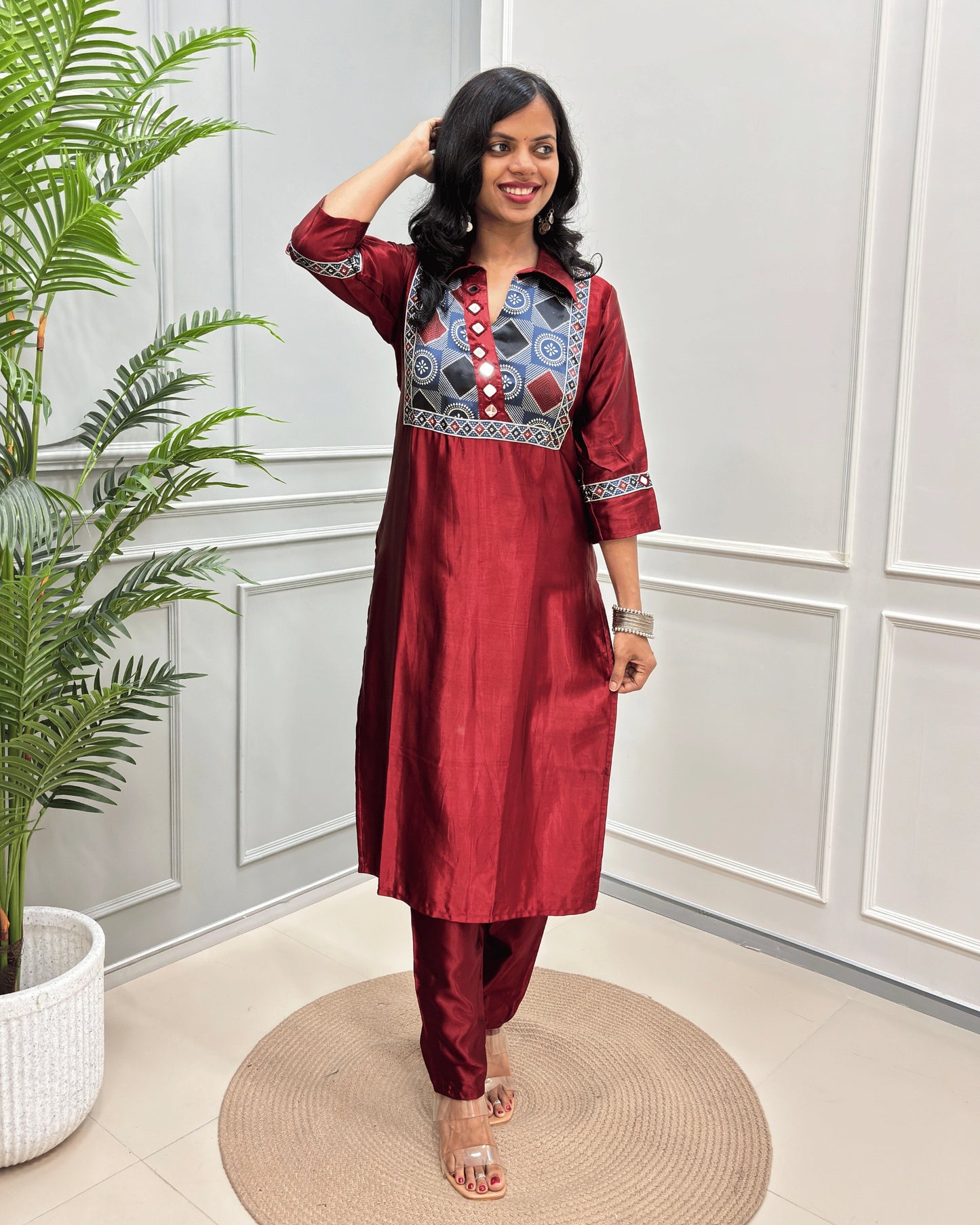 Red Women's Ajrakh Mashru Silk Long Kurti with colorful embroidered detailing on the chest and sleeves, paired with black pants, standing in a decorated room with a potted plant and paneled wall.