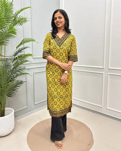 Mustard Yellow Women's Ajrakh Hand-block Pure Cotton Long-Kurti with yellow and Black geometric patterns, paired with Black Flared pants, standing on a round woven mat in front of a white paneled wall with a green potted plant.