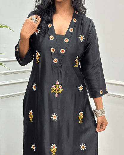 Close-up view of Black Mashru Silk Long Kurti with colorful embroidered floral motifs and a Hand-mirror work on V-neckline, Front & Cuffs, Full Sleeves ,  standing against a light gray paneled wall. 