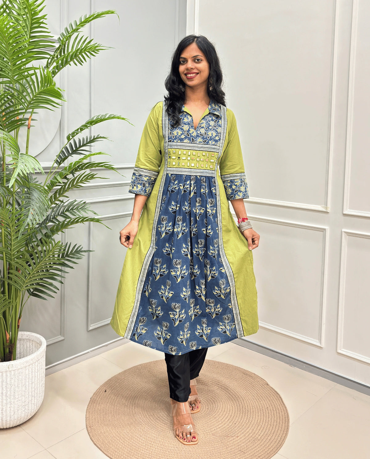 Green Women Ajrakh Vegetable Dyed Plant Based Rayon Cotton Long Kurti Featured with Green & Blue Floral print , Aline pattern with side slight, paired with black pants, standing on a round woven mat in front of a white paneled wall with a potted plant on the left.
