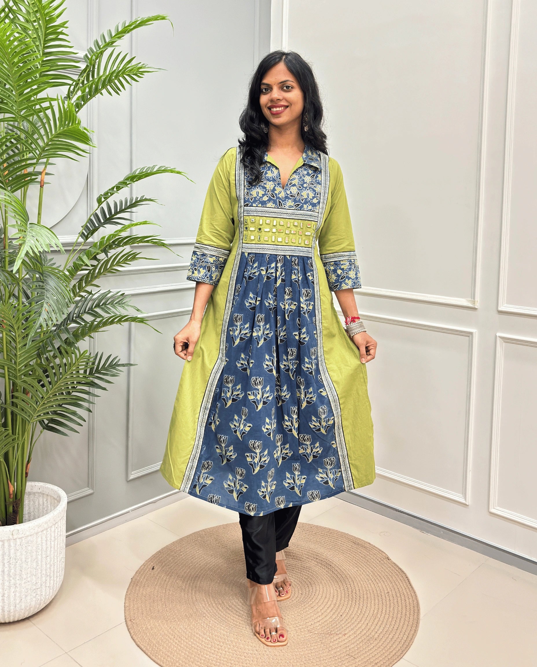 Green Women Ajrakh Vegetable Dyed Plant Based Rayon Cotton Long Kurti Featured with Green & Blue Floral print , Aline pattern with side slight, paired with black pants, standing on a round woven mat in front of a white paneled wall with a potted plant on the left.