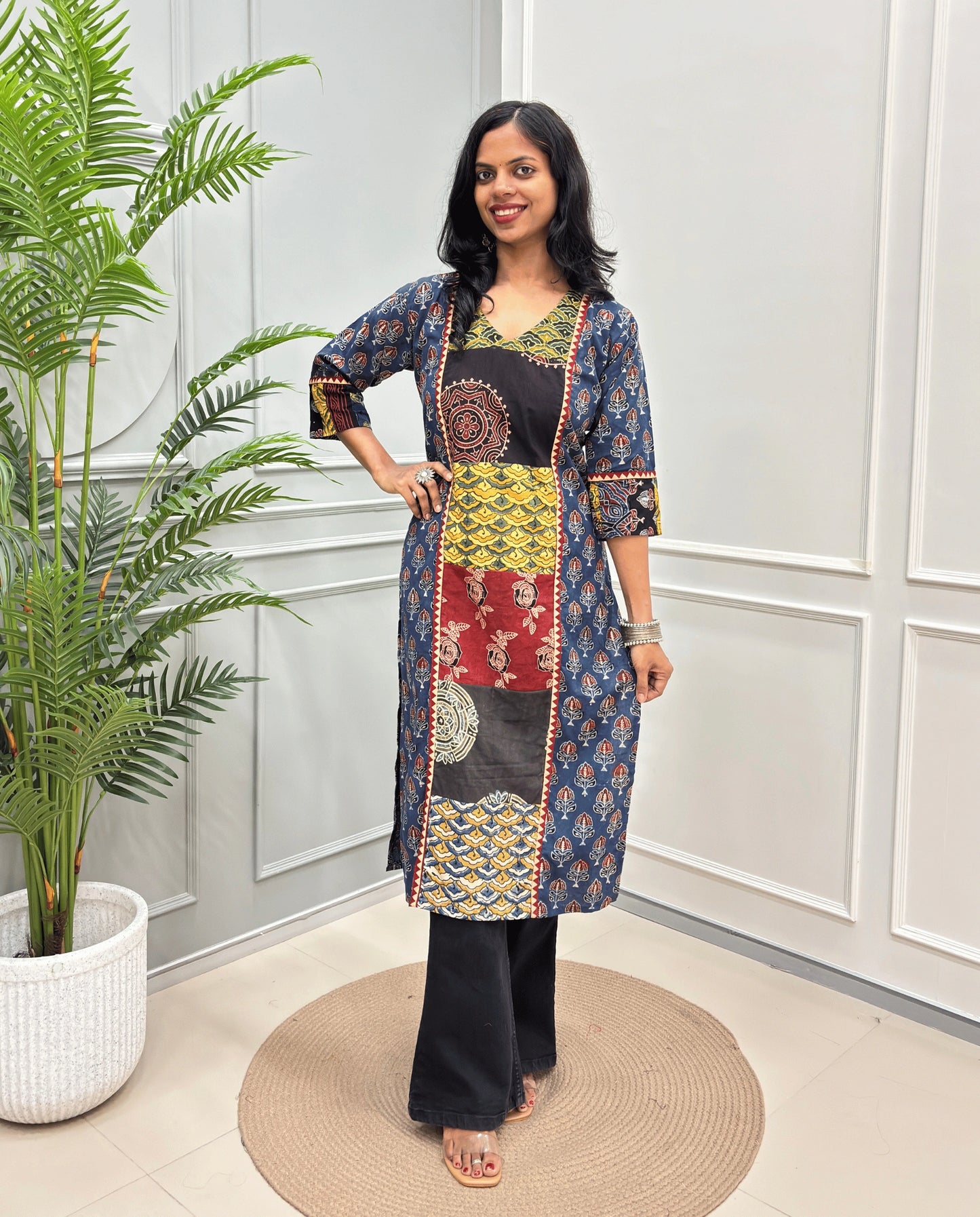 Blue women's Hand-block Ajrakh  pure cotton Long Kurti with three-quarter sleeves, Ajrakh print on front and sleeves and black wide-leg pants, standing on a round woven mat in front of a white paneled wall next to a potted palm.
