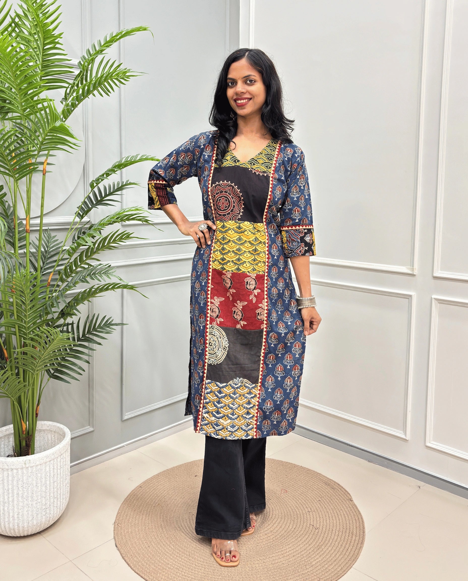 Blue women's Hand-block Ajrakh  pure cotton Long Kurti with three-quarter sleeves, Ajrakh print on front and sleeves and black wide-leg pants, standing on a round woven mat in front of a white paneled wall next to a potted palm.