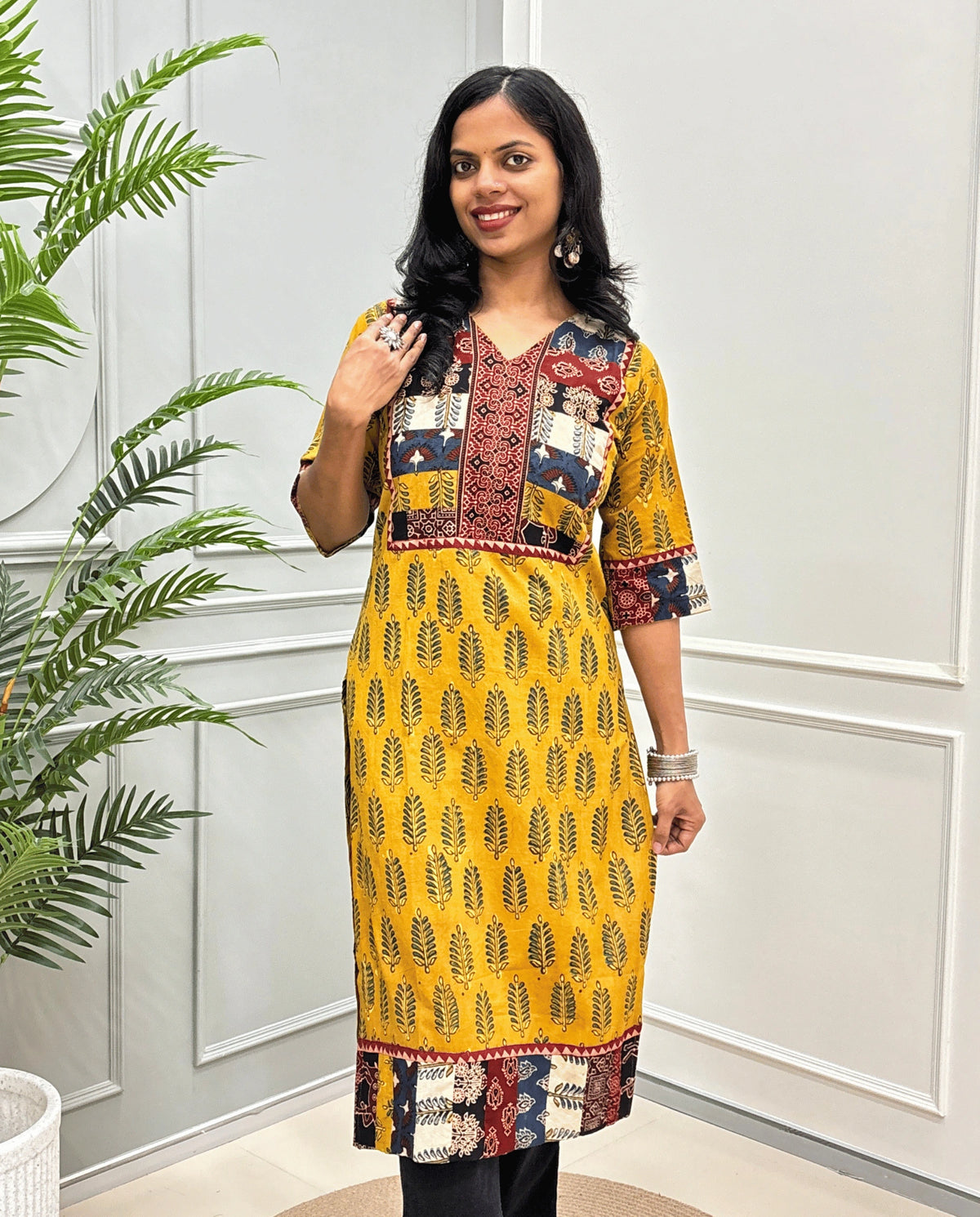 Yellow Women's Hand-block Ajrakh Pure cotton Long-Kurti with multicolor patchwork-style yoke on Front, Cuffs and hem, V-shaped neck, Ajrakh print on front & Sleeves, paired with black Flared pants, standing indoors against a white paneled wall next to a potted plant.