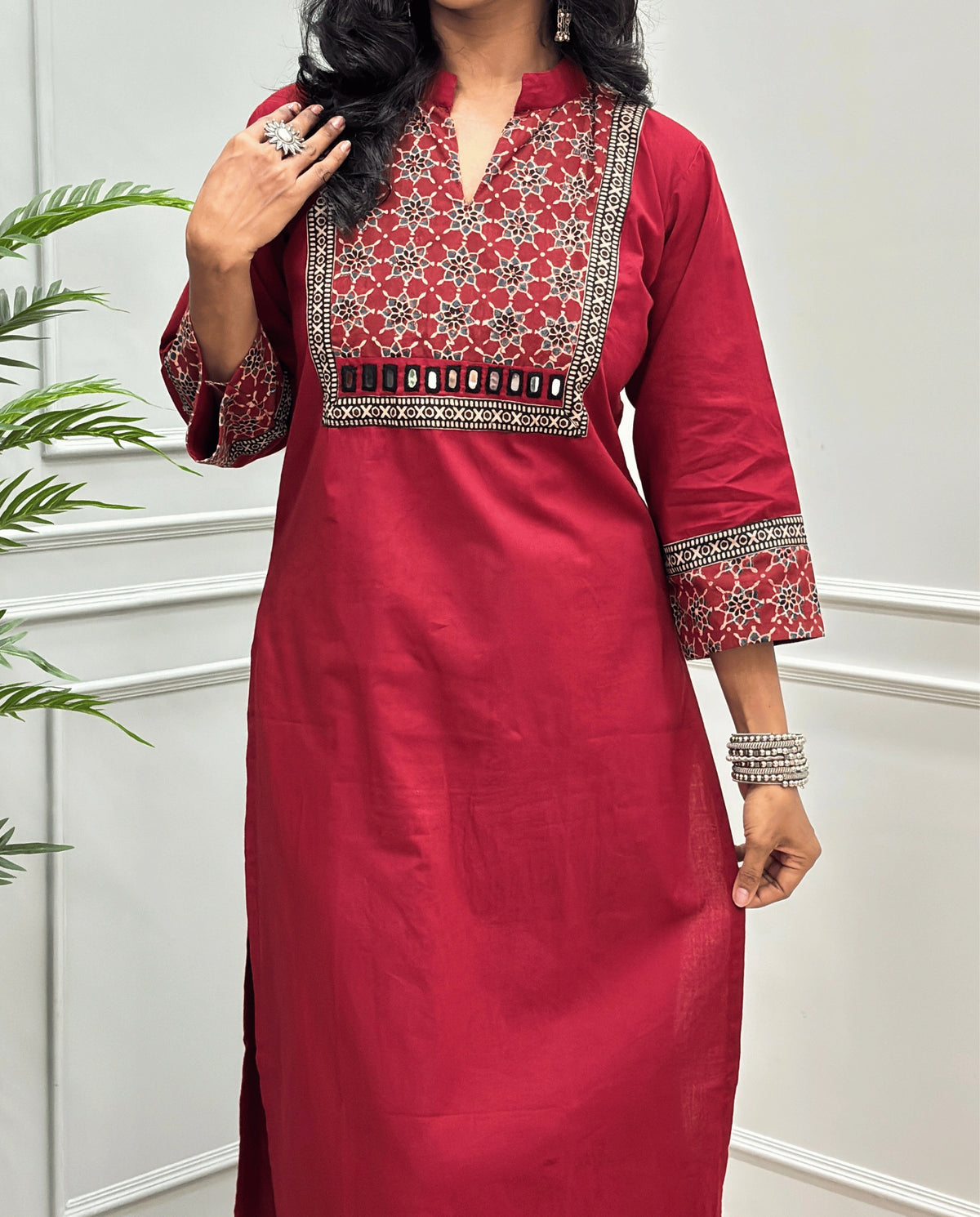 Close-up of Red Hand-block Ajrakh Pure cotton Long kurti with three quarter sleeves with printed cuffs,  intricate black & beige geometric embroidery, with hand mirror work and thread work on border of mirror , with Mandarian collar V-neck and sleeve cuffs, standing against a light gray paneled wall.