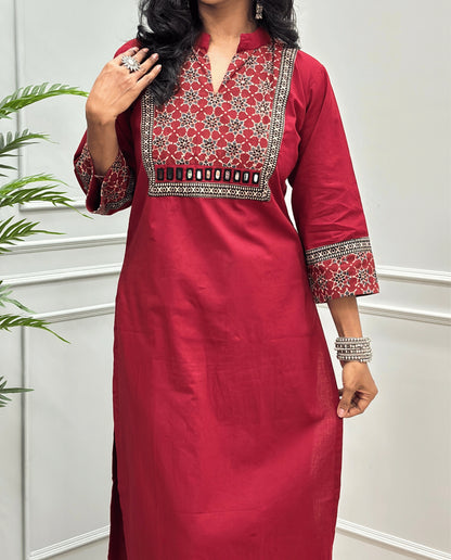 Close-up of Red Hand-block Ajrakh Pure cotton Long kurti with three quarter sleeves with printed cuffs,  intricate black & beige geometric embroidery, with hand mirror work and thread work on border of mirror , with Mandarian collar V-neck and sleeve cuffs, standing against a light gray paneled wall.