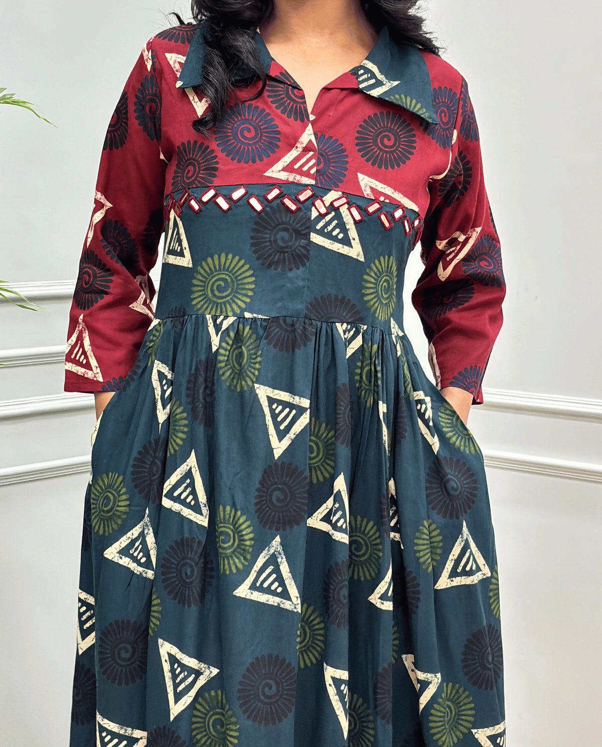 Bottle-Green Women's Ajrakh Vegetable dyed plant based Cotton Long-Kurti Featured with Three-Quarter Sleeves with a Red neck and cuffs, and a Green-colored collar & dress featuring a geometric pattern of triangles and spiral in Green, black, and blue, Hand-mirror work on front, standing on a round woven rug in front of a white paneled wall with a green potted plant on the left.