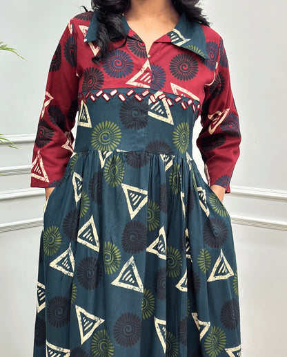 Bottle-Green Women's Ajrakh Vegetable dyed plant based Cotton Long-Kurti Featured with Three-Quarter Sleeves with a Red neck and cuffs, and a Green-colored collar & dress featuring a geometric pattern of triangles and spiral in Green, black, and blue, Hand-mirror work on front, standing on a round woven rug in front of a white paneled wall with a green potted plant on the left.