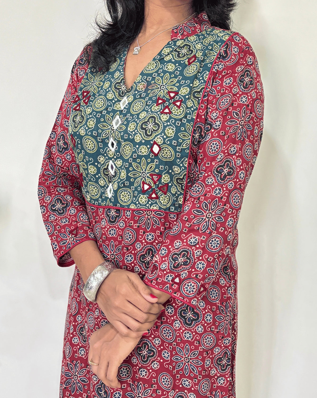 Close-up of Women's Red Ajrakh Vegetable Dyed plant based cotton long kurti Featured with full sleeves, with green, black, and red patterns, Hand-mirror work on front & Sleeves , with Mandarian V-shaped neck, paired with black flared pants, standing in a room with a white wall and a potted plant.