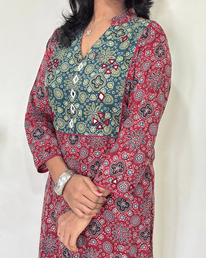 Close-up of Women's Red Ajrakh Vegetable Dyed plant based cotton long kurti Featured with full sleeves, with green, black, and red patterns, Hand-mirror work on front & Sleeves , with Mandarian V-shaped neck, paired with black flared pants, standing in a room with a white wall and a potted plant.
