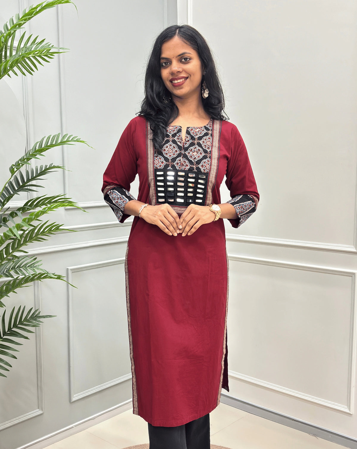 Front View of Red Women's Hand-block Ajrakh Pure Cotton Long Kurti with a red base and abstract Black, Red  & Beige geometric embroidery on the chest and sleeve cuffs, Hand-mirror work on Front, Ajrakh print border on Sides & Front  paired with black Flared pants, standing in a decorated room with a potted plant.