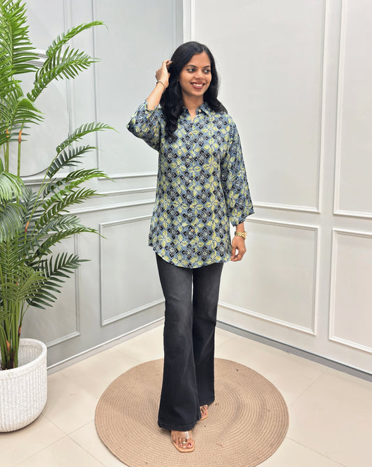 Green Women's Ajrakh Pure Modal Silk Shirt with a Floral print , black flared pants, standing on a round woven rug in front of a white paneled wall with a potted plant on the left.