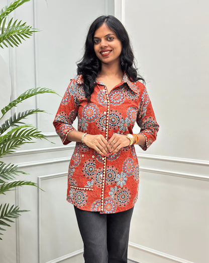 Front View of Orange women's Ajrakh pure cotton Shirt with a orange base and Multi color floral print, Hand-mirrorwork on front button style, Sleeves cuffs & pocket, paired with black flared pants. Standing on a round woven rug against a light paneled wall with a potted plant at left.