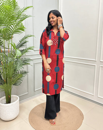 Red Women's Ajrakh Vegetable dyed plant based Cotton Long-Kurti with a geometric pattern of navy blue, beige, and black circles, paired with black Flared pant, standing in a room with a potted plant and paneled walls.