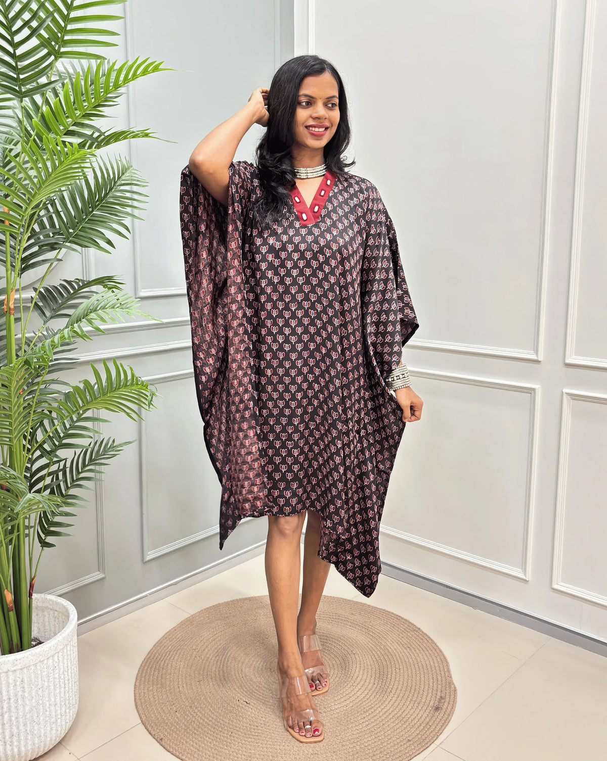 Black Women's Ajrakh Pure Modal Silk Knee Length Kaftan with a black Floral print , standing on a round woven rug in front of a white paneled wall next to a potted plant.