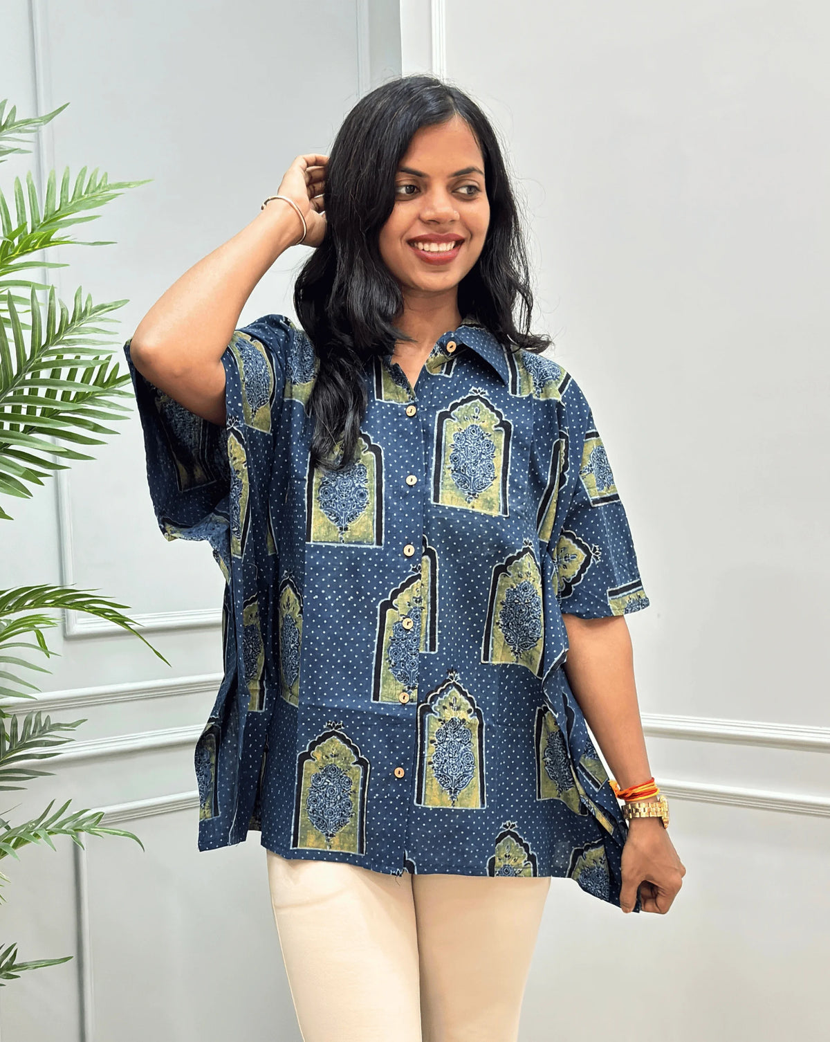 Front View of Blue Women's Ajrakh Pure Cotton Short Kaftan Shirt with a Black, Green & Yellow Floral Block Print,  Wooden Button on front, paired with beige flared pants, standing on a round woven rug in front of a white paneled wall with a potted plant.