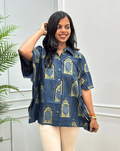 Front View of Blue Women's Ajrakh Pure Cotton Short Kaftan Shirt with a Black, Green & Yellow Floral Block Print,  Wooden Button on front, paired with beige flared pants, standing on a round woven rug in front of a white paneled wall with a potted plant.