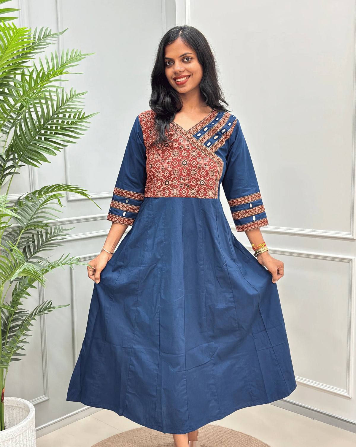Front view of Navy-blue Women's Ajrakh Pure Cotton A-line Anarkali with a Red base & Black floral yoke patterned along with Real hand-mirror work on Front & sleeve cuffs , standing on a round woven rug in front of a light gray paneled wall.