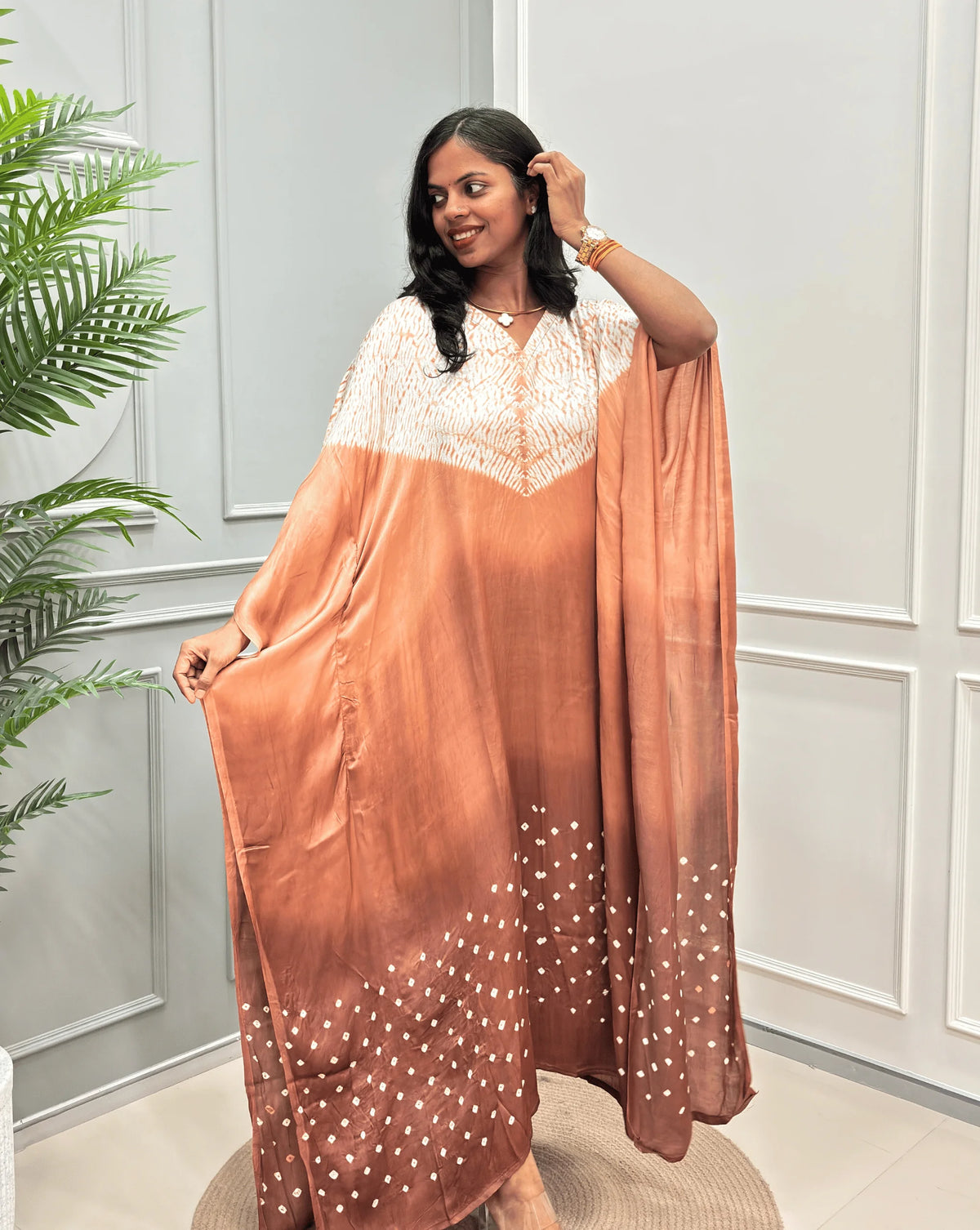 Front-Side View of Brown Women's Ajrakh Pure Modal Silk Shibori Kaftan Featured with Long-length, with a 3 shades of brown with ajrakh print on chest & Bandhani Small dots with white on Bottom, standing on a round woven rug in front of a white paneled wall.