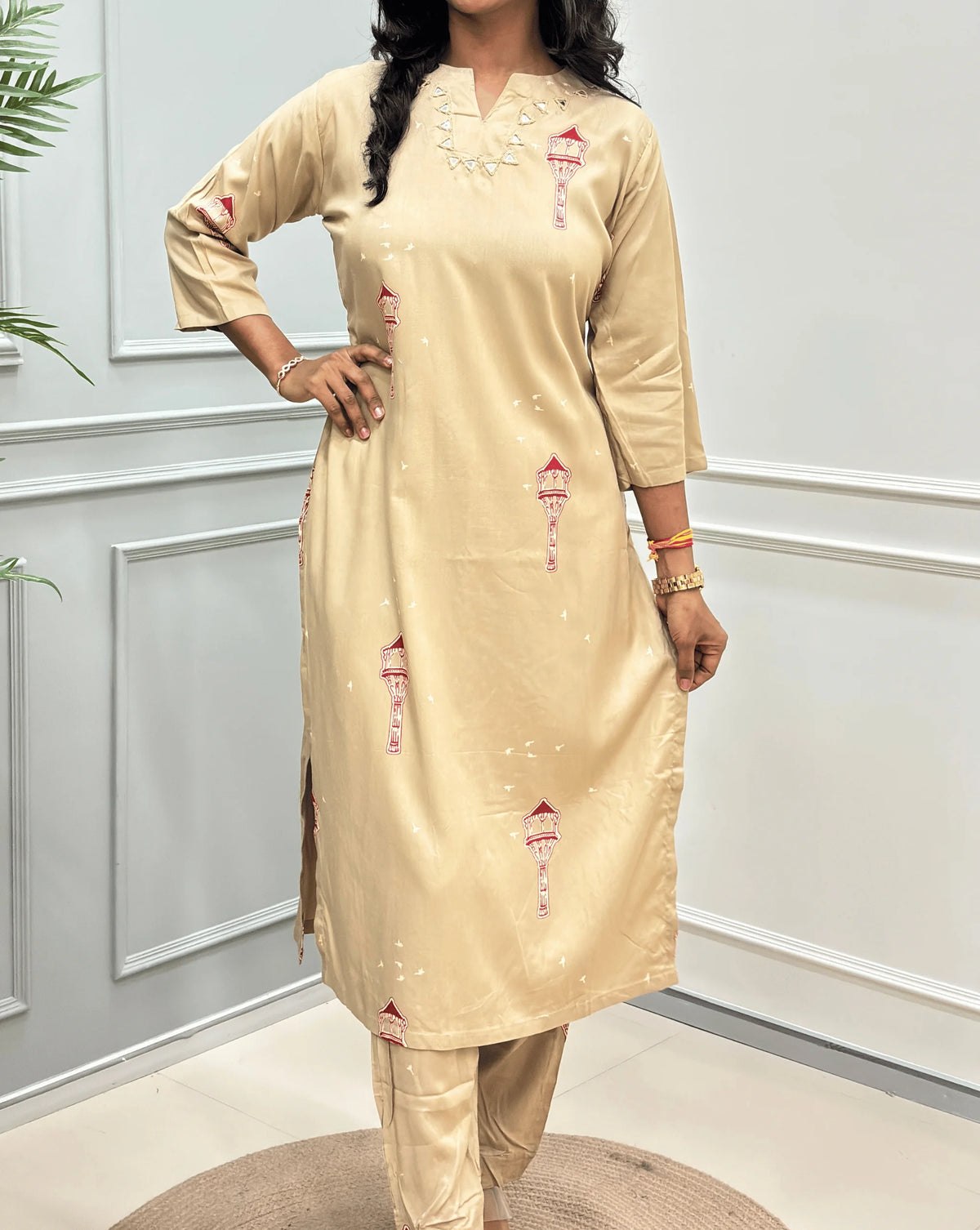 Close-up View of Beige Women's Ajrakh  Vegetable Dyed Cotton Long Co-Ord set Featured with Round V-neck & Three -Quarter Sleeves with small red and green embroidered details & Real Hand-Mirror Work on Neck-line, along with matching pants, standing in a modern room with white paneled walls and a potted plant