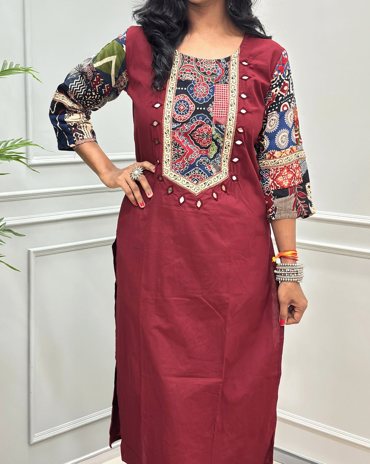 Close-up of Brick-Red women's Ajrakh Vegetable dyed plant based Cotton Co-Ord set Featured with Round neck & Three-quarter sleeves with Multi-colour ajrakh Print detailed embroidered on neckline & Sleeves with beige strip border on work , Hand-mirror work out-line on front, paired with matching teal pants, standing on a round woven mat in a bright room with a potted plant.
