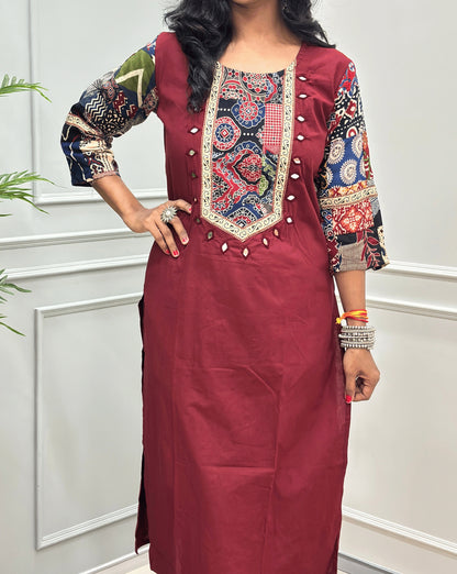 Close-up of Brick-Red women's Ajrakh Vegetable dyed plant based Cotton Co-Ord set Featured with Round neck & Three-quarter sleeves with Multi-colour ajrakh Print detailed embroidered on neckline & Sleeves with beige strip border on work , Hand-mirror work out-line on front, paired with matching teal pants, standing on a round woven mat in a bright room with a potted plant.