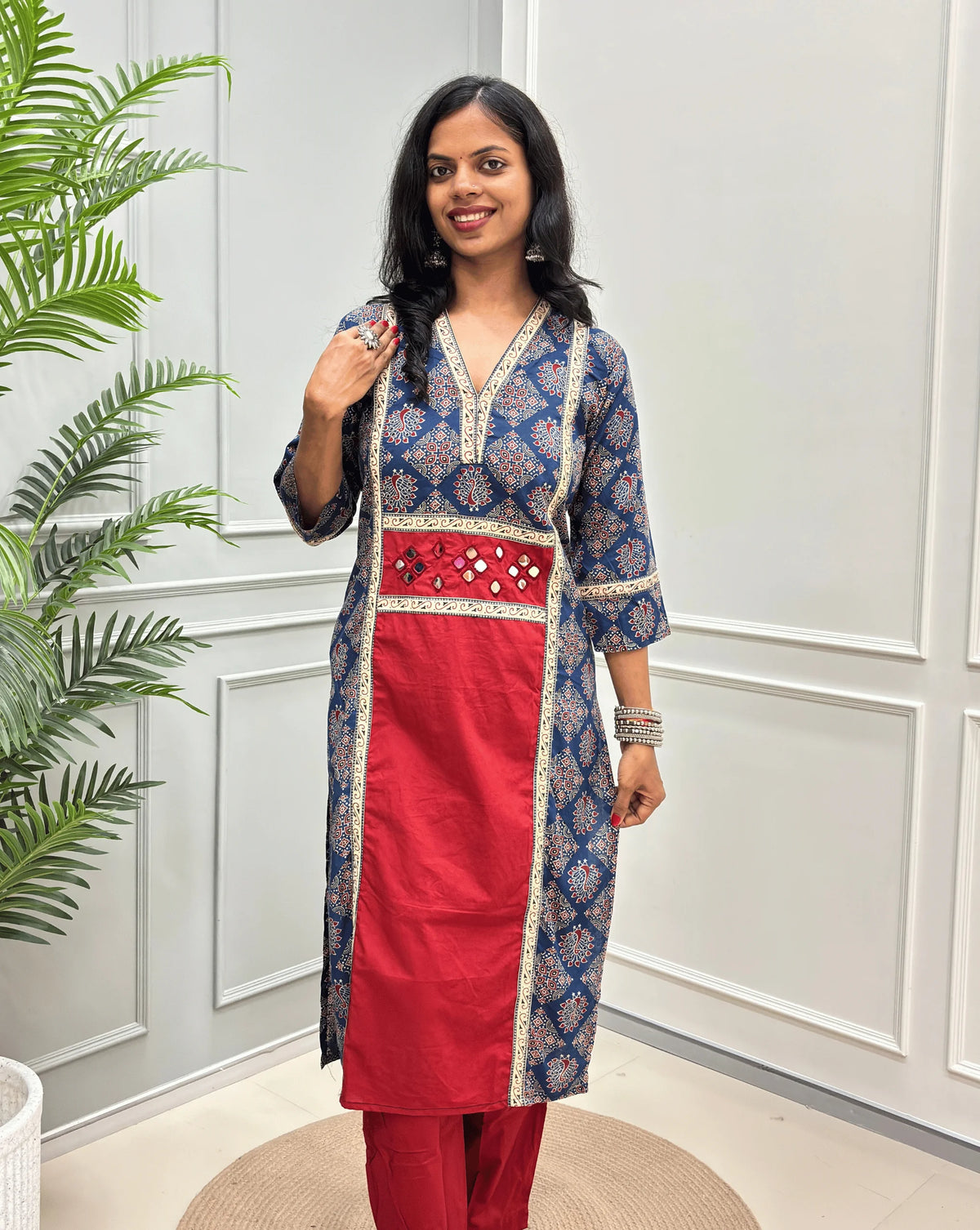 Front-View of Red Women's Ajrakh pure cotton 2 peice Co-Ord set with a Blue base & an Peacock print on Front Chest to Side & Sleeves, Hand-mirror work on Front as Belt, standing on a round woven mat in front of a white paneled wall with a potted plant.