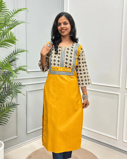 Front view of Mustard Yellow Women's Hand-block Ajrakh Pure Cotton Long Kurti with a Beige base , an abstract of Blue & Black Floral Print yoke on front & Full sleeves, Hand-mirror work on Front, paired with blue Pant, standing on a round woven mat in front of a white paneled wall.