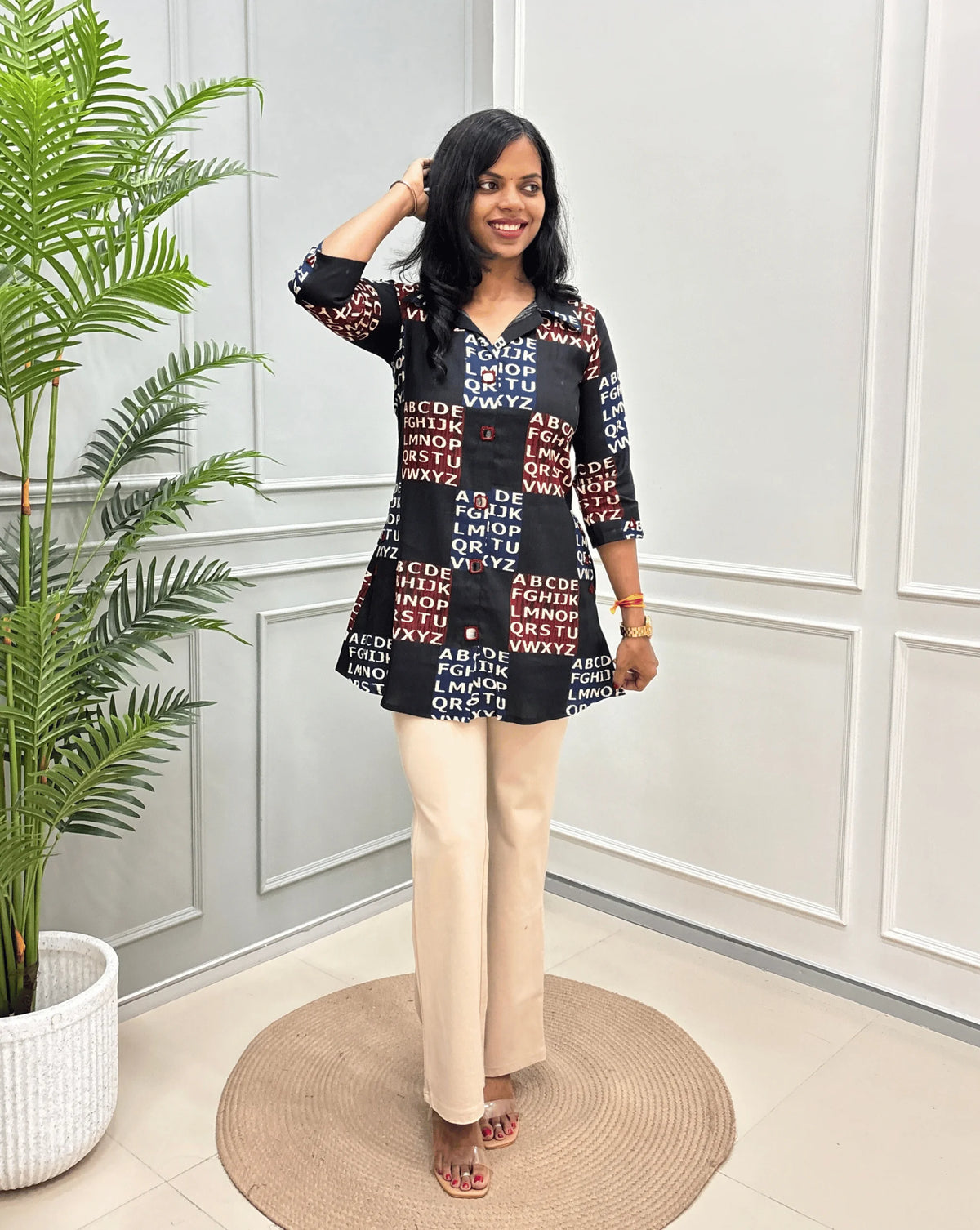 Black Women's Ajrakh Vegetable Dyed Plant based Cotton Short kurti with a Red & Blue Base, & Abstract Beige color alphabet print, paired with beige trousers, standing on a round woven rug in front of a white paneled wall with a potted plant.