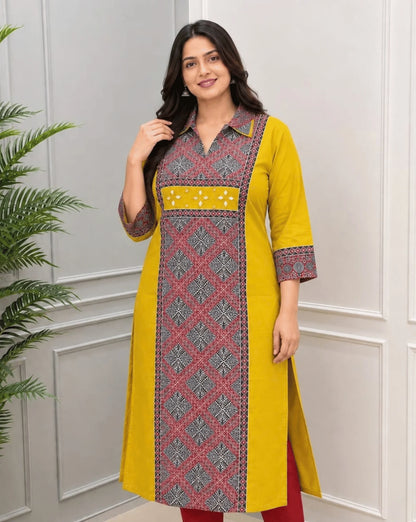 Front-Side View of Mustard Yellow Women's Ajrakh Vegetable Dyed Plant Based Cotton Long-Kurti with a Red Base,  an Abstract of Black & Beige geometric patterned panel on front & Sleeves Cuffs, Real Hand-Mirror Patch work on Front, paired with Red pants, standing in a bright room with a white & Grey paneled wall, on left side a potted plant.