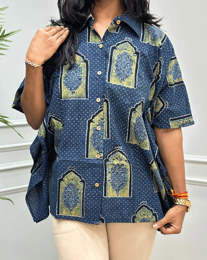 Close-up View of Blue Women's Ajrakh Pure Cotton Short Kaftan Shirt Featured with Shirt Collar neck with a Black, Green & Yellow Floral Block Print,  Wooden Button on front, paired with beige flared pants, standing on a round woven rug in front of a white paneled wall with a potted plant.