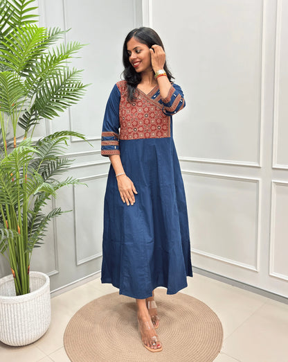 Front view of Navy-blue Women's Ajrakh Pure Cotton A-line Anarkali with a Red base & Black floral yoke patterned along with Real hand-mirror work on Front & sleeve cuffs , standing on a round woven rug in front of a light gray paneled wall.