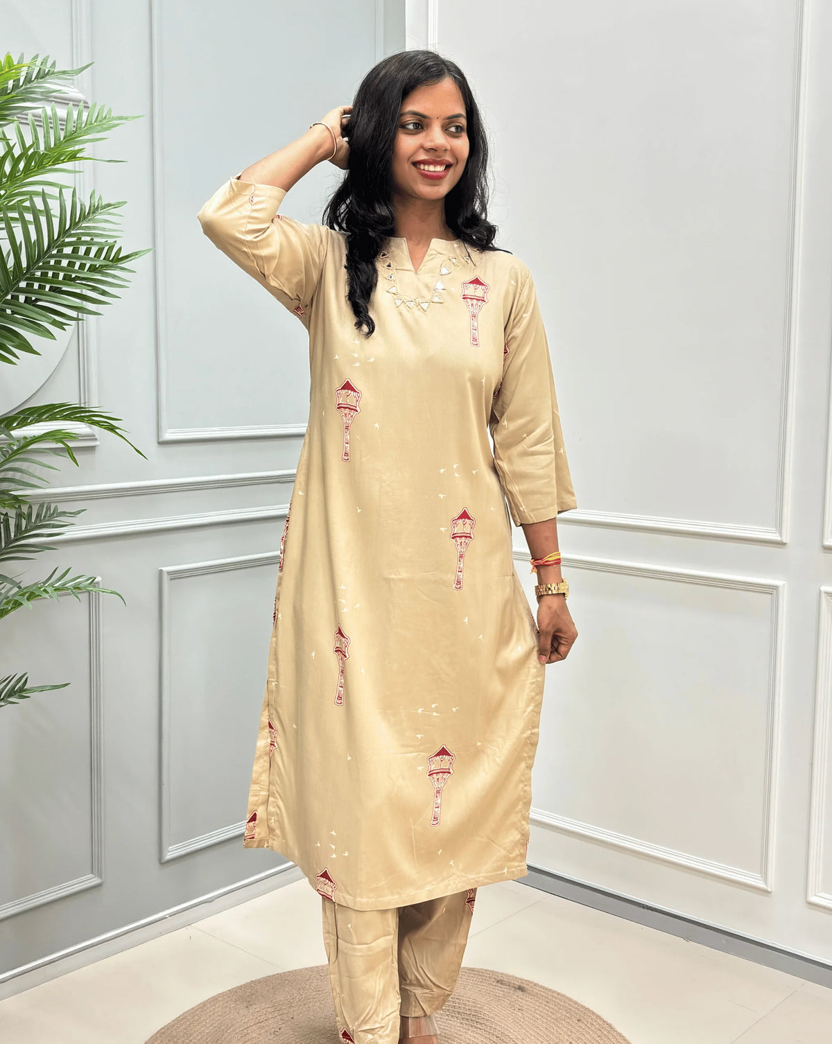 Front-View of Beige Women's Ajrakh  Vegetable Dyed Cotton Long Co-Ord set with small red and green embroidered details & Real Hand-Mirror Work on Neck-line, along with matching pants, standing in a modern room with white paneled walls and a potted plant