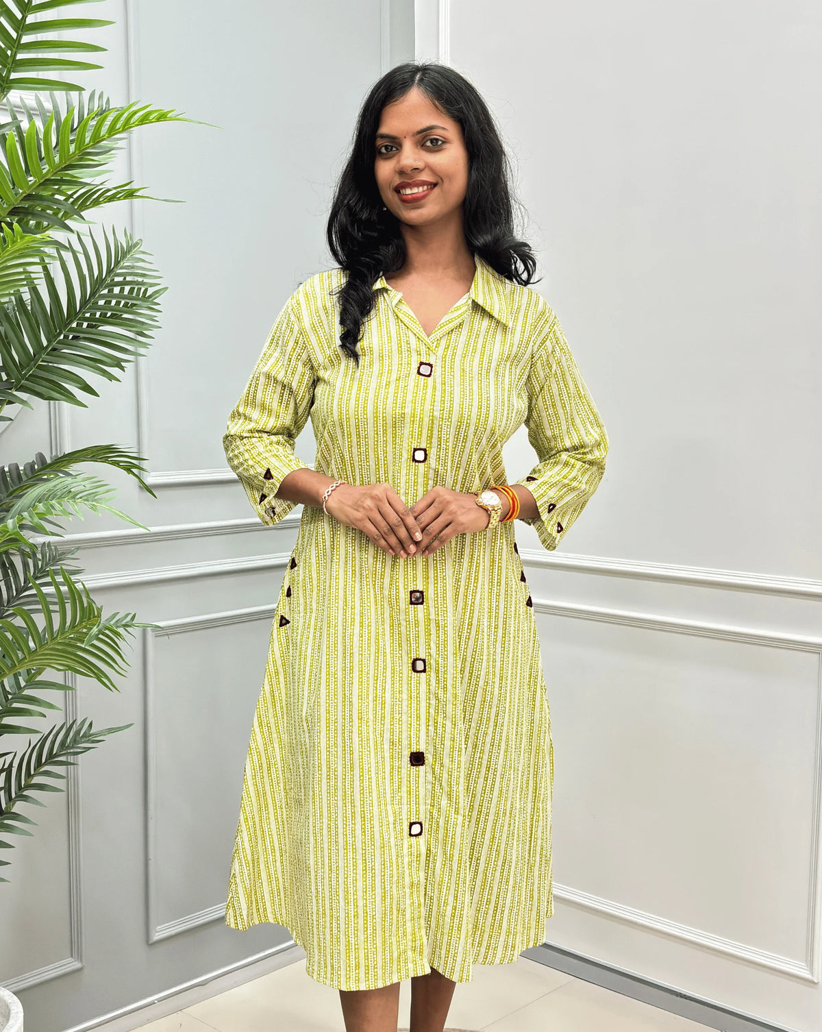 Front-View of Green Women's Ajrakh A-line Pure Cotton Long Kurti with an Ajrakh Strip Print on abstract of Green, White,  Real hand-mirror work on front & Sleeves, Along with 2 side pockets, standing on a round woven rug in front of a light gray paneled wall with a potted plant.