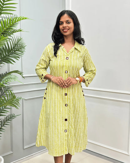 Front-View of Green Women's Ajrakh A-line Pure Cotton Long Kurti with an Ajrakh Strip Print on abstract of Green, White,  Real hand-mirror work on front & Sleeves, Along with 2 side pockets, standing on a round woven rug in front of a light gray paneled wall with a potted plant.